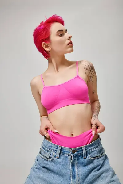 Donna elegante tatuata con capelli rosa in posa in crop top e tirando mutandine da jeans su grigio — Foto stock