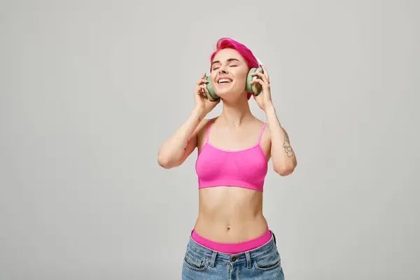 Mujer joven y feliz con el pelo rosa escuchando música en auriculares inalámbricos verdes sobre fondo gris - foto de stock