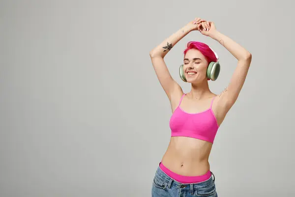 Glückliche Frau mit rosa Haaren, die Musik in drahtlosen Kopfhörern auf grauem Hintergrund hört, erhobene Hände — Stockfoto