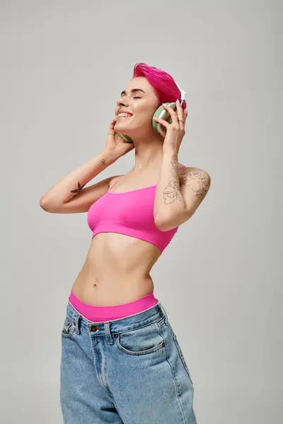 Mujer elegante tatuada con pelo rosa escuchando música en auriculares inalámbricos sobre fondo gris - foto de stock