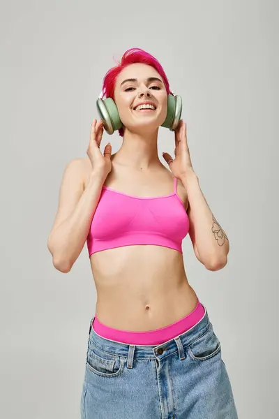 Alegre mujer perforada con pelo rosa escuchando música en auriculares inalámbricos sobre fondo gris - foto de stock