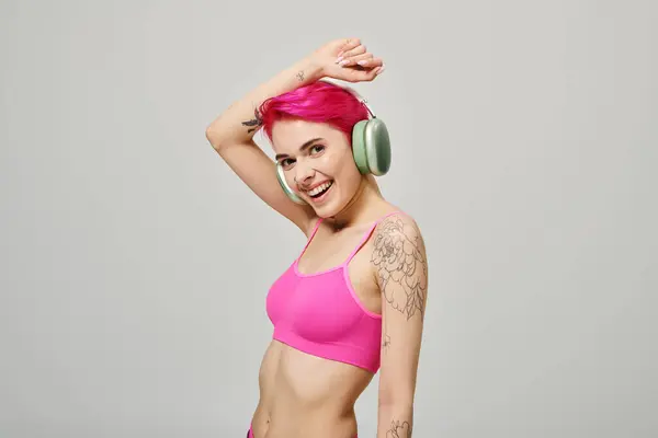Fröhliche und gepiercte Frau mit pinkfarbenen Haaren, die in drahtlosen Kopfhörern vor grauem Hintergrund Musik hört — Stockfoto