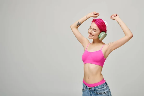 Mulher alegre e perfurada com cabelo rosa ouvindo música em fones de ouvido sem fio no fundo cinza — Fotografia de Stock