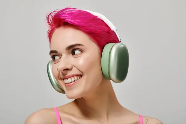 Retrato de mujer alegre perforada con pelo rosa escuchando música en auriculares inalámbricos en gris - foto de stock