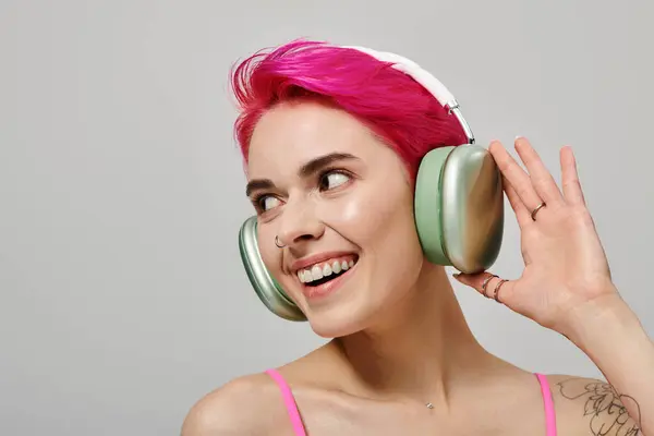 Retrato de mujer tatuada alegre con pelo rosa escuchando música en auriculares inalámbricos en gris - foto de stock