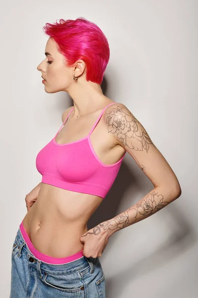 Вид збоку татуйованої молодої жінки з рожевим волоссям, позує у верхній частині культури і джинси на сірому тлі — стокове фото