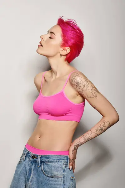 Портрет татуйованої та експресивної жінки з рожевим волоссям, що позує у верхній частині культури та джинси на сірому — стокове фото