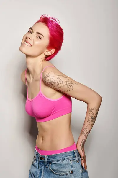Mulher tatuada e expressiva com cabelo rosa posando com as mãos em bolsos de jeans em cinza — Fotografia de Stock