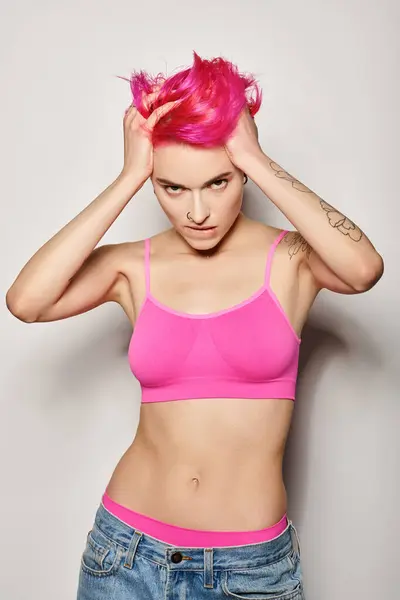 Татуированная и выразительная женщина трогает свои розовые волосы и позирует в кукурузном топе на сером фоне — стоковое фото