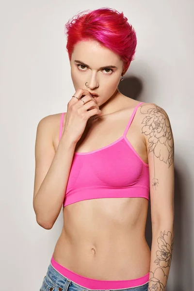 Татуированная и пронзительная женщина с розовыми волосами и позирующая в ярком топе и джинсах на сером фоне — стоковое фото