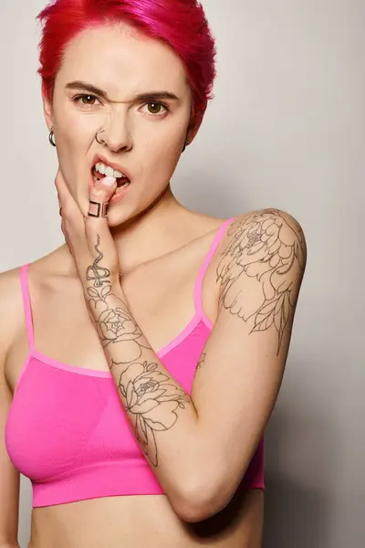 Татуированная и пронзительная женщина с розовыми волосами и позирующая в кусающем пальце на сером фоне — стоковое фото
