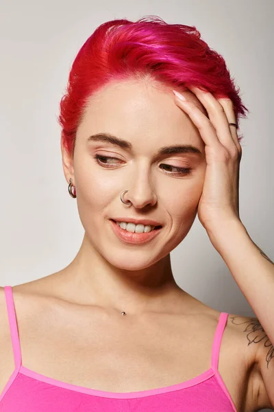 Portrait of beautiful and positive young woman touching pink hair and looking away on grey backdrop — Stock Photo