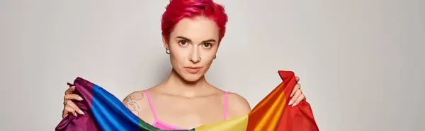 Портрет татуйованої молодої жінки з рожевим волоссям позує з веселкою lgbt прапор на сірому, банер — стокове фото