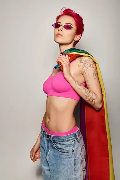 Mujer joven con pelo rosa y gafas de sol posando con bandera de arco iris lgbt sobre fondo gris, orgullo - foto de stock