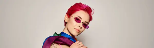 Young woman with pink hair and stylish sunglasses posing with lgbt rainbow flag on grey, banner — Stock Photo