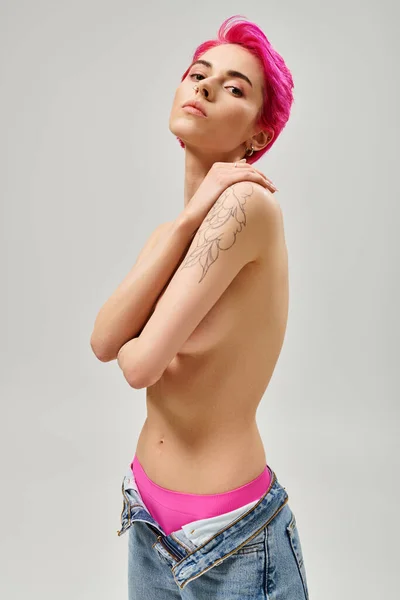 Mujer joven tatuada y en topless con el pelo rosa cubriendo los pechos y posando sobre fondo gris - foto de stock