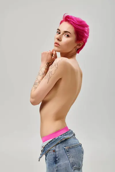 Tattooed and topless young woman with pink hair covering breasts and posing on grey backdrop — Stock Photo