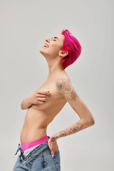 Tattooed and topless young woman with pink hair covering breasts and smiling on grey backdrop — Stock Photo