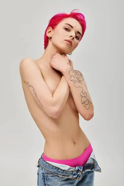 Portrait de jeune femme tatouée et seins nus avec des cheveux roses couvrant les seins sur fond gris — Photo de stock