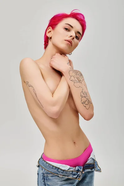 Portrait of tattooed and topless young woman with pink hair covering breasts on grey background — Stock Photo