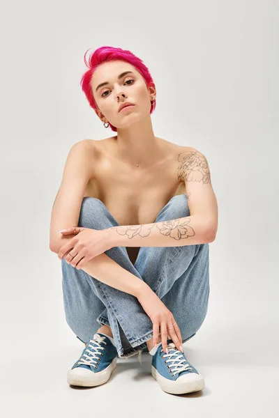 Mujer tatuada y en topless con el pelo rosa sentado en vaqueros vaqueros azules sobre fondo gris, sexy - foto de stock