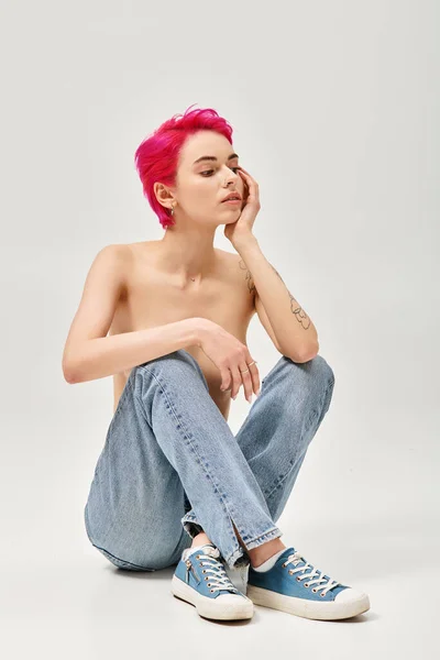 Tattooed and topless woman with pink hair sitting in denim jeans on grey background, relaxed pose — Stock Photo