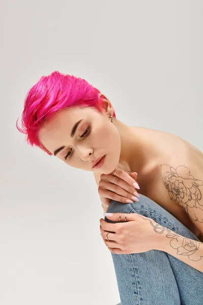 Tattooed and topless woman with pink hair sitting in denim jeans on grey background, dreamy — Stock Photo