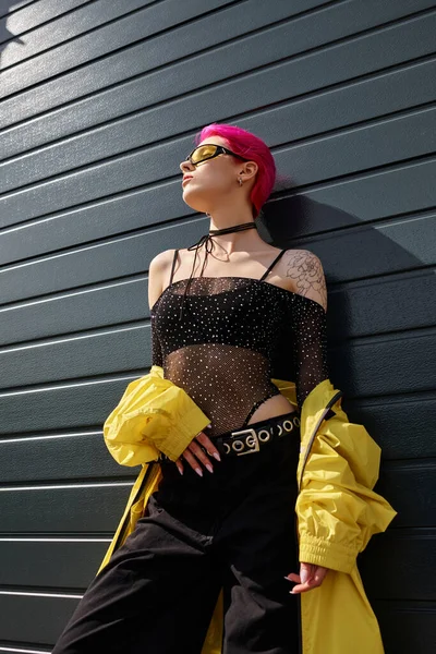 Pink haired young woman in yellow sunglasses and stylish outfit posing outdoors, low angle view — Stock Photo