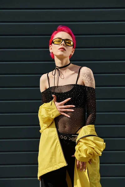 Pink haired young woman in yellow sunglasses and stylish outfit posing on urban street outdoors — Stock Photo