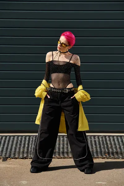 Pink haired young woman in sunglasses and stylish attire posing with hands in pockets on street — Stock Photo