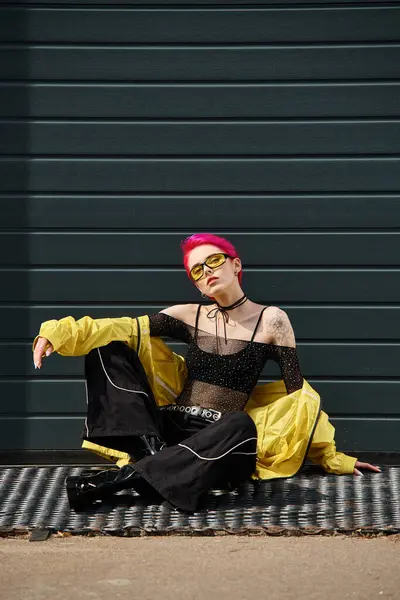Pink haired young woman in yellow sunglasses and stylish attire sitting on urban street outdoors — Stock Photo