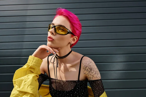 Nachdenkliche junge Frau mit rosa Haaren und Tätowierung posiert mit Sonnenbrille und trendiger Streetwear auf der Straße — Stockfoto