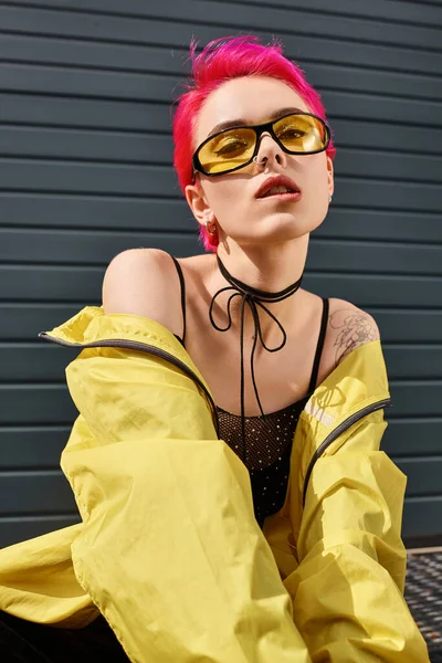 Rose jeune femme aux lunettes de soleil jaunes et tenue élégante lèvre mordante et regardant la caméra — Photo de stock