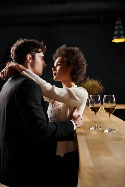 Cariñosa pareja interracial abrazando cerca de copas de vino en el mostrador del bar durante la fecha en el restaurante - foto de stock