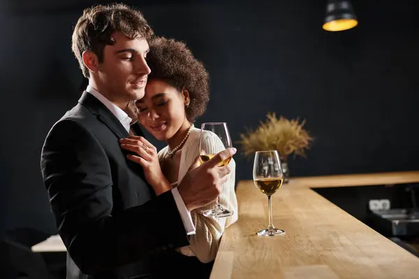 Happy man in suit holding glass of wine and hugging curly african american girlfriend during date — Stock Photo