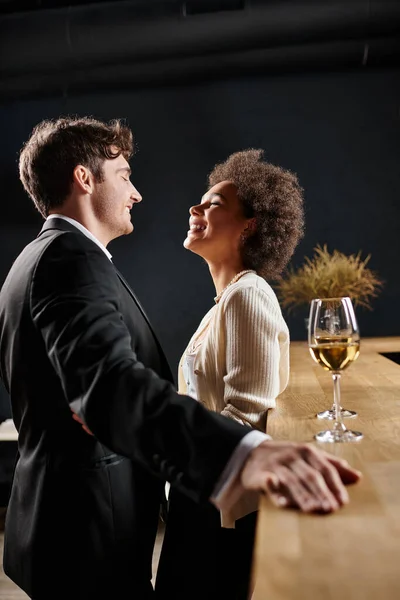 Happy multicultural couple laughing while standing near bar counter with wine glasses during date — Stock Photo