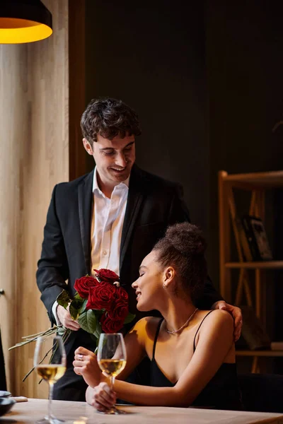 Красивый мужчина в костюме держит розы возле счастливой африканской американки в ресторане на День Святого Валентина — стоковое фото