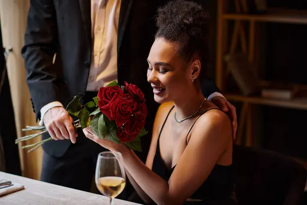 Uomo in giacca e cravatta che tiene rose vicino felice giovane donna africana americana nel ristorante a San Valentino — Foto stock
