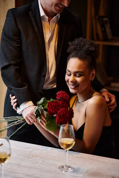 Мужчина в костюме держит розы возле счастливой африканской женщины в ресторане на День Святого Валентина — стоковое фото