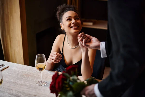 Hombre de traje cogido de la mano de la mujer afroamericana feliz en el restaurante el día de San Valentín - foto de stock