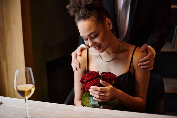 Счастливая африканская американка с красными розами рядом с мужчиной в костюме, стоящим позади нее во время свидания — стоковое фото