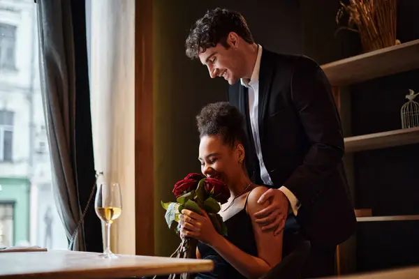 Довольная африканская американка, нюхающая красные розы рядом с мужчиной в костюме, стоящим позади нее во время свидания — стоковое фото