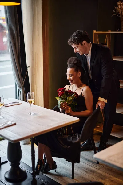 Джентльмен, стоящий за стулом счастливой африканской американки, держащей розы во время свидания — стоковое фото