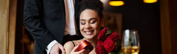 Mann schenkt herzförmige Schachtel an glückliche afrikanisch-amerikanische Frau mit roten Rosen am 14. Februar, Banner — Stockfoto