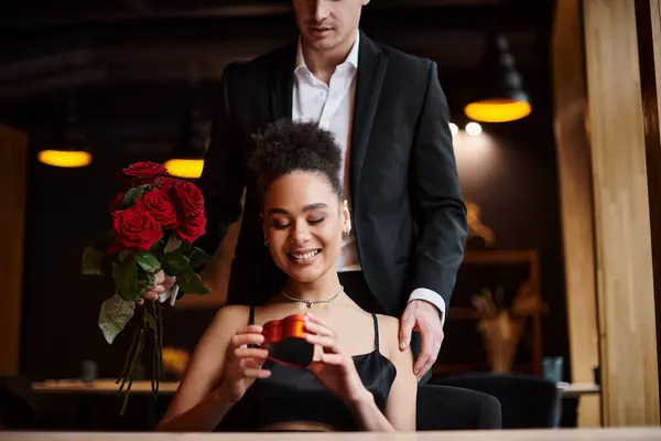 Мужчина дарит красные розы счастливой африканской женщине, держащей коробку в форме сердца в день Святого Валентина — стоковое фото