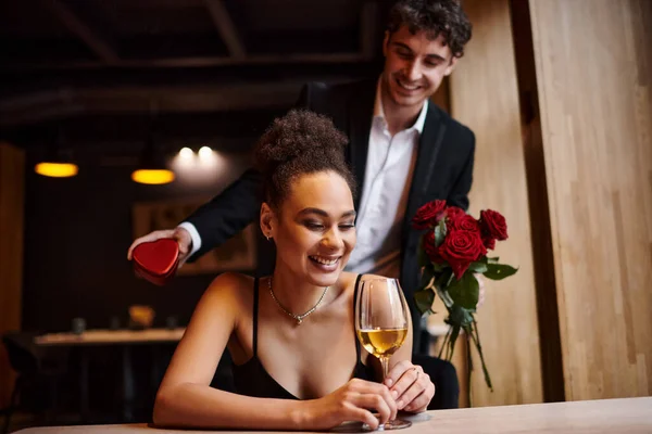 Homem segurando rosas vermelhas e caixa em forma de coração perto de mulher americana africana feliz no dia dos namorados — Fotografia de Stock