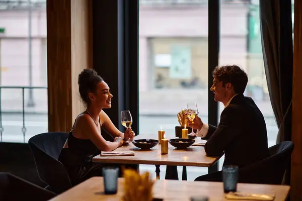 Feliz casal interracial em trajes elegantes segurando copos com vinho durante a data no restaurante — Fotografia de Stock