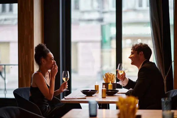 Feliz pareja interracial en elegante atuendo sosteniendo copas de vino durante la cita en el restaurante, risa - foto de stock