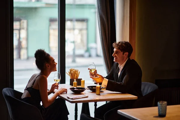 Alegre interracial casal no elegante traje segurando copos com vinho durante data no restaurante — Fotografia de Stock