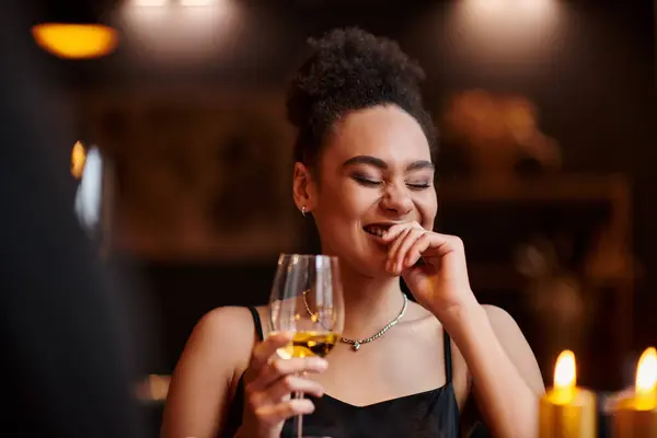 Curly african american woman laughing and holding glass of wine during date on Valentines day — Stock Photo